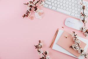Spring flat lay top view home office workspace - modern keyboard and notebook with cherry blossom branches on a pink desk background photo