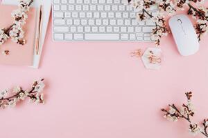 Spring flat lay top view home office workspace - modern keyboard and notebook with cherry blossom branches on a pink desk background photo