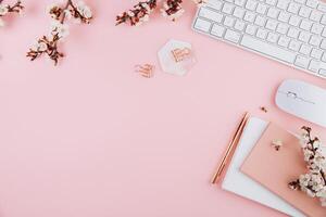 Spring flat lay top view home office workspace - modern keyboard and notebook with cherry blossom branches on a pink desk background photo