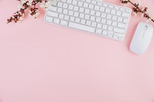 Spring flat lay top view home office workspace - modern keyboard and notebook with cherry blossom branches on a pink desk background photo