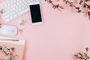 Spring flat lay top view home office workspace - modern keyboard, smartphone and notebook with cherry blossom branches on a pink desk background photo