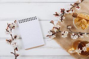 Spring composition. A cup of herbal tea, a jar of honey and cherry blossom near notebook on a white wooden background, top view. Place for text. Healthy food planning concept. photo