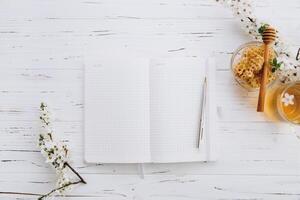 Spring composition. A cup of herbal tea, a jar of honey and cherry blossom near notebook on a white wooden background, top view. Place for text. Healthy food planning concept. photo
