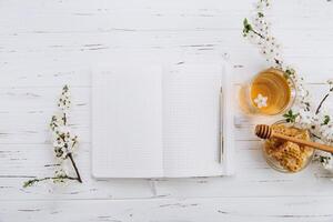Spring composition. A cup of herbal tea, a jar of honey and cherry blossom near notebook on a white wooden background, top view. Place for text. Healthy food planning concept. photo