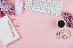 Spring flat lay top view home office workspace - modern keyboard, cup of coffee and notebook with lilac flowers on a pink desk background photo