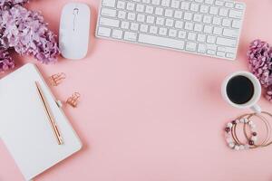 Spring flat lay top view home office workspace - modern keyboard, cup of coffee and notebook with lilac flowers on a pink desk background photo