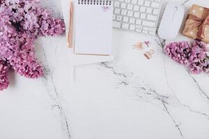 Spring flat lay top view home office workspace - modern keyboard, gift and notebook with lilac flowers on a marble desk background photo