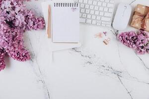 Spring flat lay top view home office workspace - modern keyboard, gift and notebook with lilac flowers on a marble desk background photo