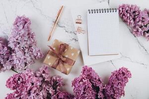 Spring flat lay top view home office workspace - gift and notebook with lilac flowers on a marble desk background photo