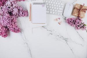 Spring flat lay top view home office workspace - modern keyboard, gift and notebook with lilac flowers on a marble desk background photo