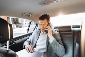 Modern businessman talking on a mobile phone and using a laptop computer while sitting in the back seat of a car. photo