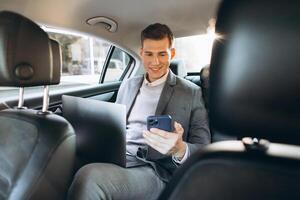 A modern businessman is talking on a mobile phone and using a tablet computer while sitting in the back seat of a car. photo