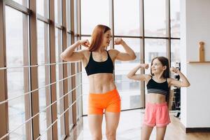 Beautiful strong woman and charming little daughter are showing their biceps and smiling while working out at gym photo