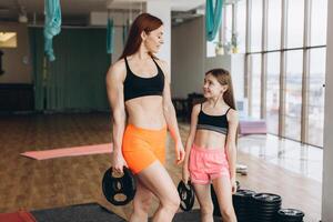 Mom and Daughter are doing exercises. Family in a gym. Little girl with strong mother are training with pancakes rods photo