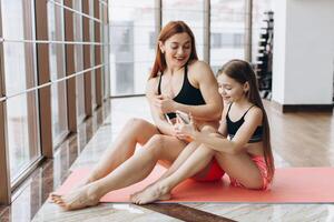 Mother and daughter relaxing after workout sitting on mat, using phone in gym for watching how do exercise photo
