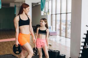 Mom and Daughter are doing exercises. Family in a gym. Little girl with strong mother are training with pancakes rods photo