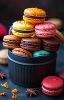 A black container filled with a variety of colorful macarons. The macarons are arranged in a pyramid shape, with the topmost one being the most colorful photo