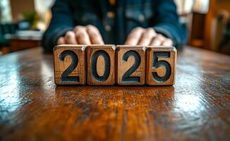 A wooden block with the numbers 2025 is on a table. The block is made of wood and has black letters on it. The block is placed on a wooden table, and it is surrounded by a person's hands photo