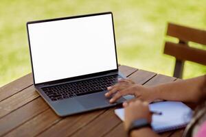 A person is seated at a wooden table, typing on a laptop under the sun, with a notepad beside them. The vibrant green grass in the background enhances the relaxing atmosphere of the workspace. photo