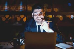 Office Manager Guy Using Voice Assistant On Smartphone Working On Laptop In Modern Office Late At Night. Selective Focus photo