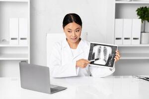 A doctor Asian lady holds a chest X-ray while explaining the results to a patient in a modern clinic. The workspace is tidy, featuring a laptop and organized files in the background. photo