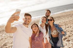 A group of five friends happily poses for selfies on the beach, capturing a joyful moment while enjoying the sun and ocean waves in the background during a perfect day. photo