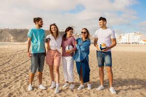 A group of five friends stand on a sandy beach enjoying each other company. They are dressed casually and are engaged in lively conversation while holding volleyballs. photo