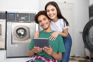 A young couple relaxes together in a laundromat, with the man seated and holding a tablet. The woman stands behind him, smiling and enjoying their time while waiting for laundry. photo