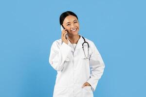 Asian doctor wearing a white coat and a stethoscope smiles while talking on the phone. The bright blue background highlights her joyful demeanor, suggesting a positive conversation or consultation. photo