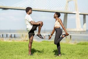 Two African American friends engage in a stretching exercise in a lush park by the river. They enjoy each other's company while preparing for a workout against a backdrop of a bridge. photo