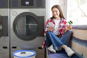 A young woman sits comfortably on a bench in a laundromat, using her smartphone while holding a payment card. She smiles as she waits for her laundry to finish. photo