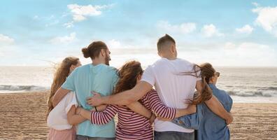A group of friends stands together on the beach, overlooking the ocean as the sun sets, with their arms around each other, capturing a moment of camaraderie and joy, back view photo