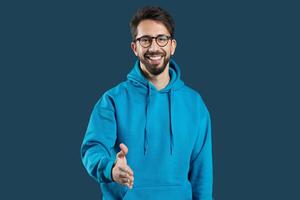A cheerful young man wearing a blue hoodie offers a handshake with a warm smile, creating an inviting and friendly atmosphere suitable for personal or professional interactions. photo
