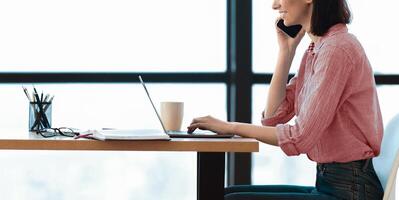 Unrecognizable girl talking on mobile phone and using laptop computer in modern office, side view, panorama, copyspace photo