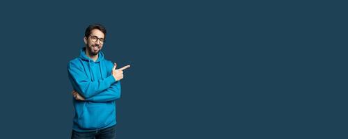 A young man wearing a blue hoodie stands against a solid dark background, smiling and pointing to the left. His casual outfit and friendly demeanor create an inviting atmosphere, copy space photo