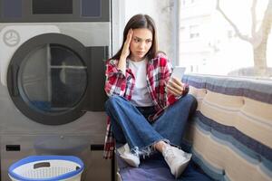 A young woman sits on a bench in a laundromat, looking stressed while checking her phone. She has a plaid shirt on and appears to be waiting for her laundry to finish. photo