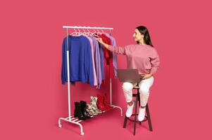 A woman sits on a stool, working on a laptop while examining clothing on a rack. The vibrant background enhances the bright colors of the apparel displayed, setting a lively atmosphere. photo