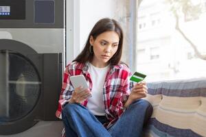 A young woman sits in a brightly lit laundromat, holding a smartphone in one hand and a credit card in the other, looking thoughtful as she prepares to make a payment. photo