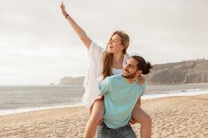 A couple shares a playful moment at the beach, with one partner giving a piggyback ride while laughing and waving. The ocean waves and soft sand create a lively, joyful atmosphere. photo