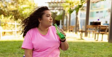 A woman sits on the grass in a park, basking in the sunlight while sipping from a green beverage. Surrounding her are trees and benches, creating a relaxing environment. photo
