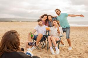 A group of five friends gathers on the beach, sitting in chairs and posing cheerfully for a photo. They hold drinks and snacks, creating a relaxed and joyful atmosphere by the shoreline. photo