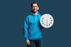 A cheerful young man wearing a blue hoodie smiles while holding a large wall clock. The background is a solid blue, creating a vibrant and lively atmosphere. photo