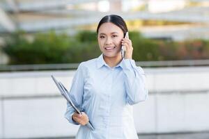 Asian woman stands outdoors, smiling as she speaks on her phone and holds a stack of documents. The background features greenery and modern architecture, suggesting a professional setting. photo