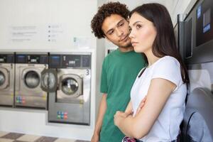 A young couple stands in a laundromat, one person looks at the other with interest while the other appears contemplative, reflecting the mood of their conversation during laundry time. photo