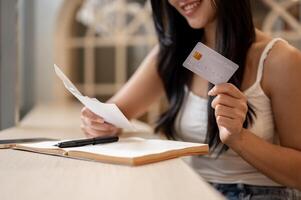 A close-up of a happy Asian woman holding a credit card and reviewing receipts at a table indoors. photo
