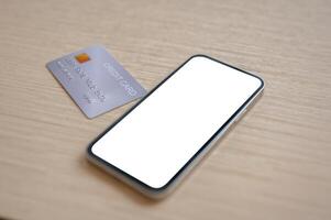 A close-up of a credit card and a smartphone with a white screen mockup placed on a wooden table indoors. photo