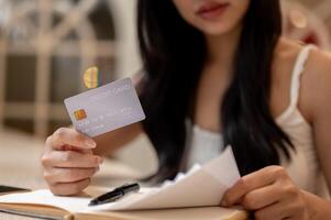 A thoughtful Asian woman holding a credit card and reviewing receipts at a table indoors. photo
