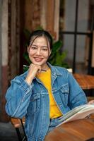 A charming, positive Asian woman in a denim jacket is reading a book while sitting in a coffee shop. photo