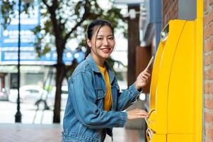 A beautiful Asian woman in a denim jacket is standing in front of an ATM, smiling at the camera. photo