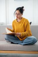 A beautiful Asian woman in casual clothes focuses on reading a book on a couch at home. photo